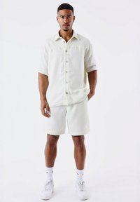 Lichtcrème kortemouwen shirt en bijpassende shorts van een gestructureerd materiaal. Met knoopsluiting aan de voorkant, gecombineerd met witte sneakers en enkelkousen.