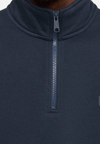 Navy fleece mikina s vysokým límcem, polovičním zipem vepředu, hladkým povrchem a malým logem na hrudi.