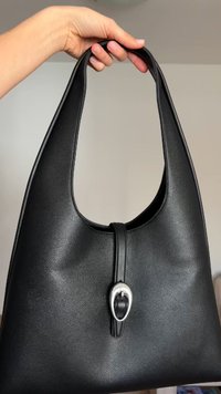 Χέρι που κρατά μια μαύρη δερμάτινη τσάντα ώμου με ασημένια πόρπη μπροστά σε απλό φόντο.