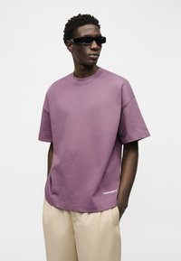 Άνδρας που φοράει μαύρα ορθογώνια γυαλιά ηλίου, φαρδιά μωβ μπλούζα Carhartt και μπεζ παντελόνι, στέκεται με τα χέρια στις τσέπες σε λευκό φόντο.