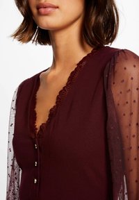 Bordeauxfarbene Bluse mit tiefem V-Ausschnitt, mit voluminöser Spitze verziert und durchsichtigen gepunkteten Ärmeln. Verfügt über drei dekorative Knöpfe entlang des Ausschnitts.