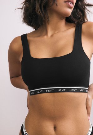 Nő fekete sportmelltartóban és hozzáillő alsóneműben, amelyeken a "NEXT" márkanév szerepel az elasztikus pántokon, a hát mögött összekulcsolt kézzel pózol.