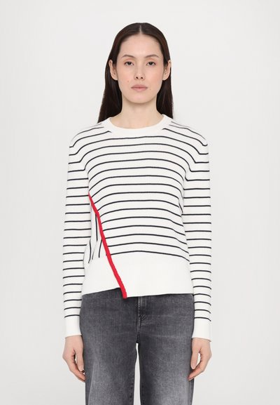 Witte trui met horizontale zwarte strepen en een diagonale rode accent. Heeft een ronde hals en geribbelde manchetten. Gecombineerd met donkergrijze jeans.