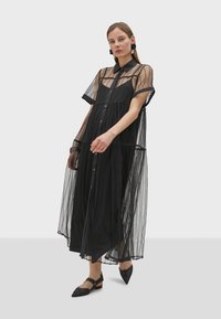Robe noire transparente avec un bouton à l'avant, manches courtes et un design à volants. Accentuée de motifs à rayures et portée par-dessus une simple robe noire.