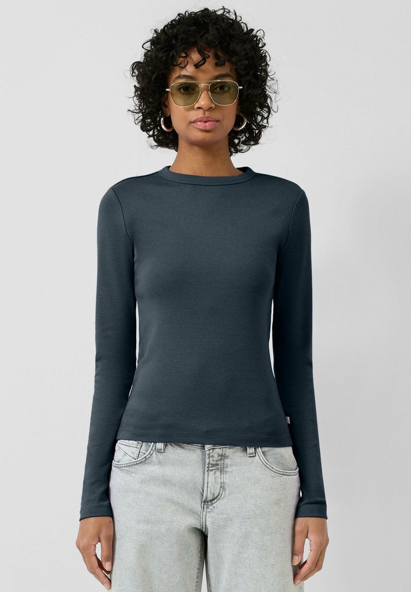 Eng anliegendes Langarmshirt in Dunkelgrau aus weichem Stoff. Es verfügt über einen runden Ausschnitt und minimalistische Nähte. Kombiniert mit hellen Jeans.