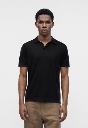 Homme debout face à nous, portant un polo noir à manches courtes et un pantalon marron clair sur un fond clair uni.