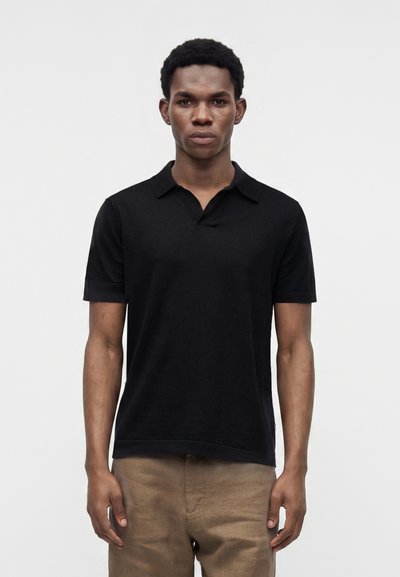 Mann, der frontal steht und ein schwarzes kurzärmeliges Poloshirt sowie hellbraune Hose vor einem schlichten hellen Hintergrund trägt.