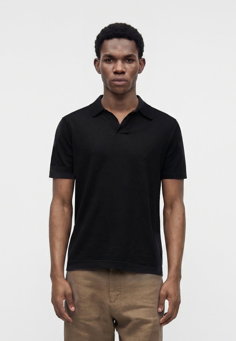 Homme debout face à nous, portant un polo noir à manches courtes et un pantalon marron clair sur un fond clair uni.