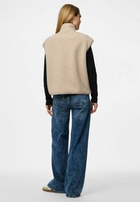 Vrouw in een beige fleece vest, zwart langarm T-shirt, wijdvallende blauwe spijkerbroek en beige sneakers, staand met haar rug naar de camera op een witte achtergrond.