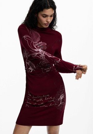 Femeie care ajustează o rochie burgundy cu mâneci lungi, cu modele florale abstracte și text scris de mână, stând în fața unui fundal alb simplu.