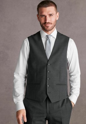 Grijze op maat gemaakte gilet met vijf knopen, voorzien van twee zijzakken, gedragen over een witte shirt en aangevuld met een lichtgrijze stropdas.