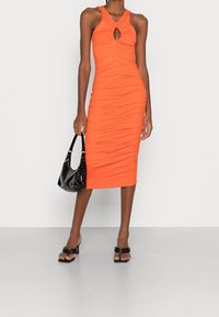 Oranžové těsně přiléhající šaty s ribbovanou texturou, klíčovou dírou v přední části a bezrukávovým designem. Kombinované s černou lakovanou kabelkou a černými sandály na blokovém podpatku.