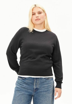 Femme blonde portant un pull noir par-dessus une chemise blanche et un jean bleu, debout avec une main dans le dos sur un fond blanc.