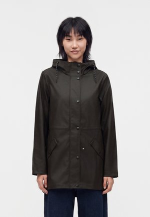 Jeune femme aux cheveux noirs portant un imperméable à capuche vert foncé avec des boutons-pression et des poches avant, debout devant un fond uni.