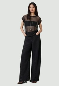 Top de malha preta com riscas horizontais, combinado com calças largas pretas com bainhas desfiadas. Modelo está de pé com as mãos nos bolsos.