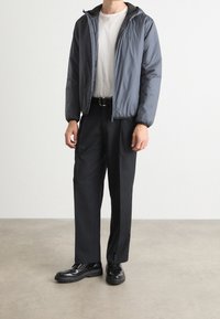 Grau gepolsterte Jacke mit Reißverschluss, mit Kapuze, kombiniert mit schwarzen maßgeschneiderten Hosen und schwarzen Schuhen mit dicken Sohlen.