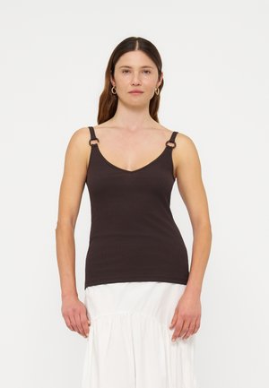 Femme aux cheveux bruns et lisses porte un haut noir sans manches avec des détails d'anneaux sur les bretelles et une jupe blanche, debout devant un fond uni.
