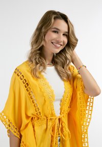 Keltainen kevyt kimono, jossa on kukkakirjontareunus, avonainen etuosa ja nyörikiinnitys. Siinä on monimutkaisia kuvioita ja virtaava muotoilu.