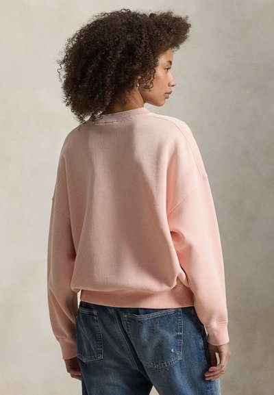 Ανοιχτό ροζ φούτερ με άνετη εφαρμογή, ριμπ σχέδιο στο λαιμό και ώμους που πέφτουν χαλαρά. Συνδυάζεται με μπλε τζιν παντελόνι με πίσω τσέπες.