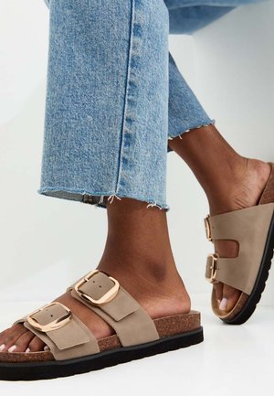 Pieds portant des sandales camel à double boucle associées à un jean en denim bleu clair coupé au-dessus de la cheville sur fond blanc.