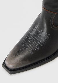 Black leather boot featuring pointed toe, contrasting stitched patterns, and decorative embroidery. Smooth texture with subtle shine.