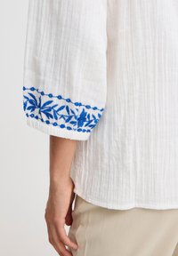 Top in tessuto bianco con strisce texturizzate; presenta un motivo floreale blu ricamato sui polsini. Vestibilità ampia, maniche a tre quarti.