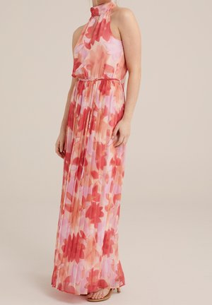 Kobieta ubrana w bezrękawnikową, wysoko zabudowaną, sięgającą podłogi plisowaną sukienkę w różowo-czerwony kwiatowy wzór z paskiem wiązanym w talii.