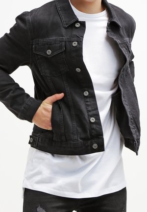 Personne portant une veste en denim noire par-dessus un t-shirt blanc, avec une main dans la poche de la veste, sur un fond blanc uni.