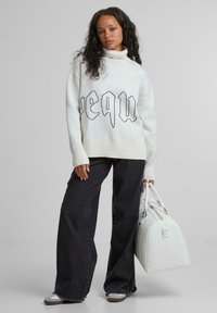 Weißer Pullover mit schwarzem Grafiktext, gerippten Bündchen und hohem Kragen; kombiniert mit weitem dunklen Jeans und einer weißen Handtasche mit Muster.