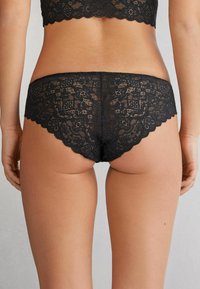 Schwarze Spitzenunterwäsche mit floralem Muster, gewellten Rändern und transparenter Textur. Das Design bietet eine tief sitzende Passform.