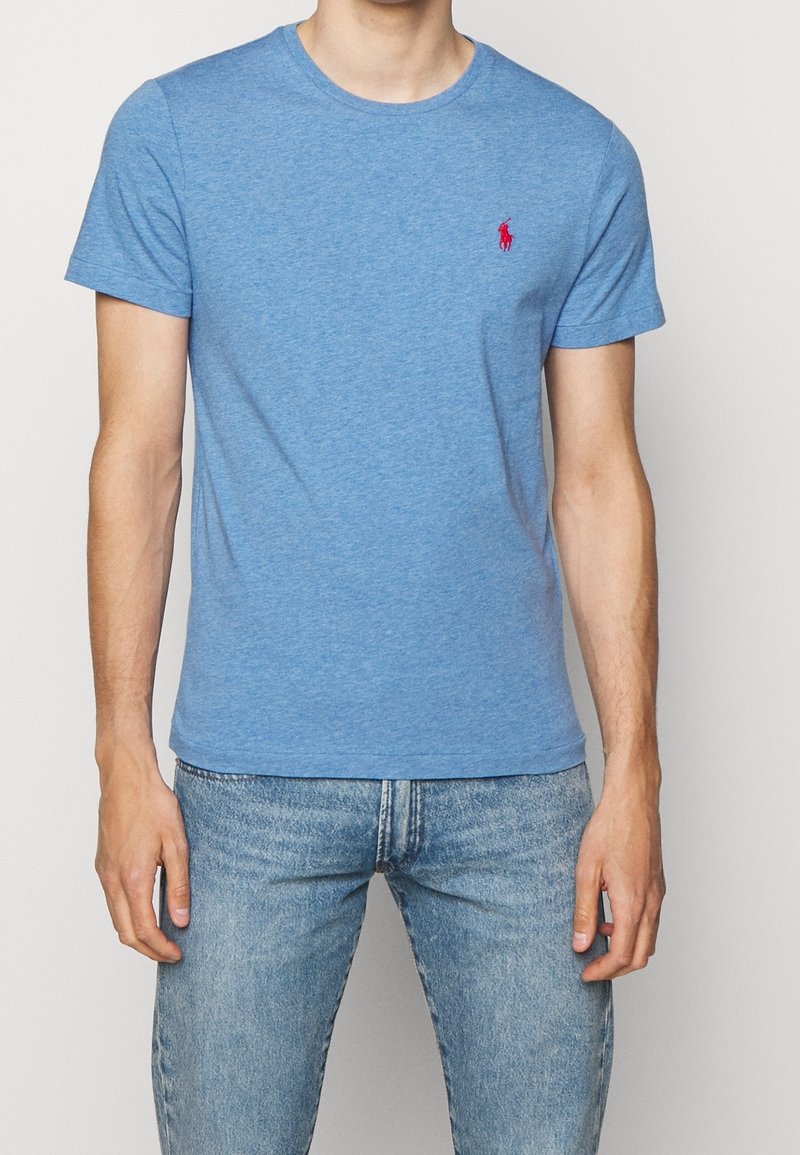 Blue cotton t-shirt with short sleeves, round neck, featuring a small red embroidered logo on the chest. Worn with light blue jeans.