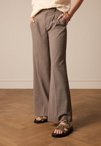 Lichtbruine broek met wijde pijpen en een plooi aan de voorkant. Gecombineerd met zwarte sandalen met een groene luipaardprintriem. Glad textuur, op maat gemaakte pasvorm.
