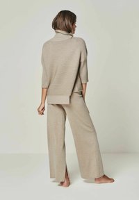 Beige Strickpullover mit Rollkragen und überschnittenen Schultern, kombiniert mit weit geschnittenen Hosen. Besitzt eine gerippte Textur und seitliche Schlitze am Saum.