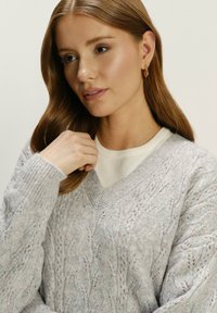 Jasnoszary sweter dziany z teksturowanymi wzorami, w serek, z prążkowanymi mankietami, nałożony na białą koszulę.