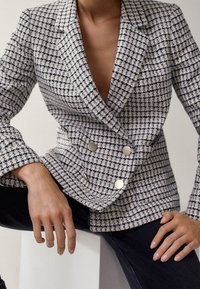 Femme portant un blazer à carreaux texturé, croisé avec des boutons argentés, associé à un pantalon noir, assise sur un bloc blanc.