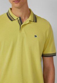 Geel poloshirt van een textuurstof, met een grijze gestreepte kraag en een logo-opdruk op de linkerkant van de borst. Korte mouwen.