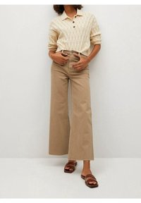 Femme portant un pull en tricot beige avec des boutons, un pantalon large taille haute beige, et des sandales à glisser marron, se tenant contre un mur uni.