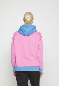 Felpa con un corpo rosa e cappuccio e orlo blu, realizzata in un materiale morbido. Il design include elementi a contrasto e polsini a costine.