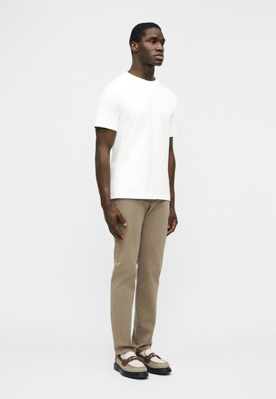 Homme debout portant un t-shirt blanc uni à manches courtes, un pantalon marron clair et des mocassins beige et marron, sur un fond uni.