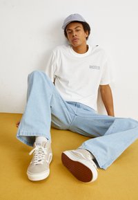 T-shirt en coton blanc avec le logo "Dickies", pantalon en denim large bleu clair, baskets beiges avec accents blancs et casquette rayée.