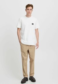 White cotton t-shirt with a square black logo on the left chest, paired with beige trousers and dark shoes, featuring a relaxed fit.
