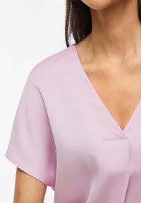 Mujer que lleva una blusa de manga corta en tono rosa claro con escote en V, mostrando el hombro, el escote y la parte superior del pecho.