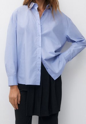 Woman wearing a loose light blue button-up shirt and black pinstriped skirt with pleats, standing with one hand on her hip.