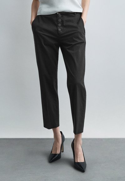 Γυναίκα που φοράει μαύρο κοντό παντελόνι με τέσσερα διακοσμητικά κουμπιά και μαύρα μυτερά παπούτσια με ψηλό τακούνι, στέκεται σε γκρι πάτωμα.