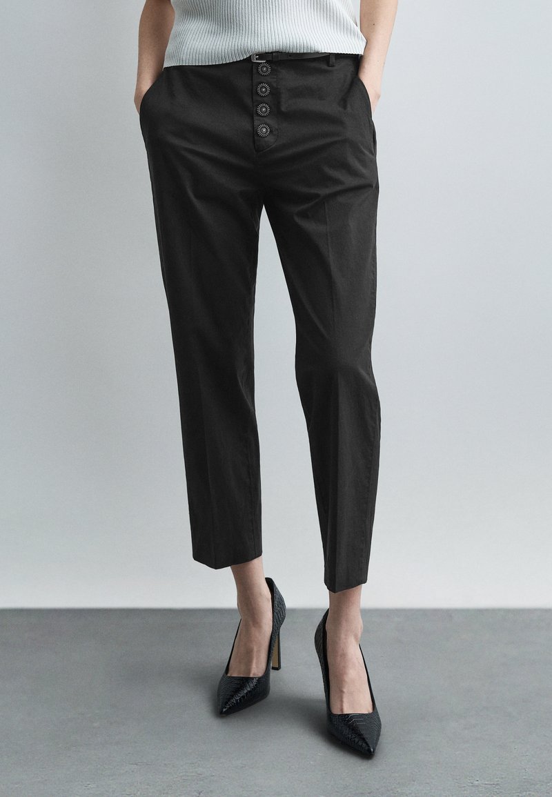 Γυναίκα που φοράει μαύρο κοντό παντελόνι με τέσσερα διακοσμητικά κουμπιά και μαύρα μυτερά παπούτσια με ψηλό τακούνι, στέκεται σε γκρι πάτωμα.