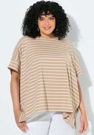 Kvinde med krøllet sort hår iført beige og hvidstribet kortærmet top og hvide bukser, smilende, stående mod en lys baggrund.