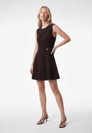 Kvinde iført en sortærmeløs kjole med guldknapper ved skuldre og talje, iført sorte sandaler med hæl og stropper.