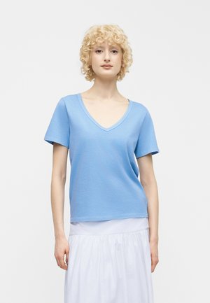 Rövid, göndör szőke hajú nő világoskék V-nyakú, rövid ujjú felsőt és fehér szoknyát viselve, egy egyszerű háttér előtt áll.