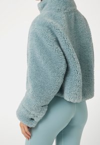 Světle modrá fleecová bunda s vysokým límcem, texturovaná měkká tkanina a široké manžety na rukávech. Střižená délka s volným střihem.