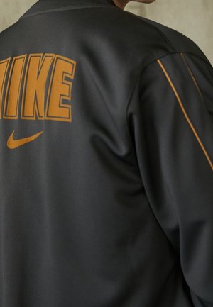 Persona con una chaqueta negra con el logo de Nike y letras naranjas en la parte trasera, con franjas naranjas en las mangas.